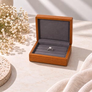 May include: A rectangular wooden ring box with a hinged lid, featuring a smooth medium-brown wood grain finish. The interior is lined with soft, dark gray velvet and contains multiple slots for rings, with a single silver-toned ring centered.