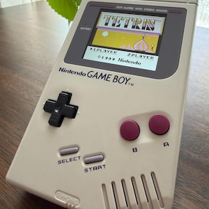 May include: A beige Nintendo Game Boy handheld console featuring a black directional pad, two purple A and B buttons, and grey Select and Start buttons. The screen displays the Tetris title screen with the text 1989 Nintendo and 1 Player 2 Player options.