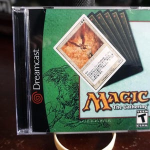 May include: A Sega Dreamcast video game case for Magic: The Gathering featuring a green background with a black dragon illustration, the Magic: The Gathering logo, a Teen ESRB rating, and a single trading card displayed over a stack of cards.