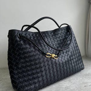 Peut inclure: Un sac cabas en cuir tressé noir doté d'une bandoulière tressée distinctive et d'un fermoir à nœud en métal doré au centre. Le sac présente une forme rectangulaire structurée avec un motif en grille cohérent sur toute la surface extérieure.