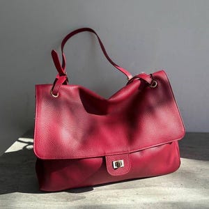 Könnte beinhalten: Eine tiefrote Handtasche mit strukturierter Oberfläche, ausgestattet mit einer großen Umschlagklappe, einem rechteckigen, goldfarbenen Drehverschluss und einem dünnen Schulterriemen, der an goldfarbenen Metallringen am oberen Rand befestigt ist.