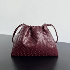 Könnte beinhalten: Eine tiefburgunderrote Handtasche mit einem geflochtenen Lederextérieur in einem markanten Gittermuster. Die Tasche verfügt über einen gerafften Verschluss im Kordelzug-Stil und zwei dünne, passende Schulterriemen aus Leder an den Seiten.