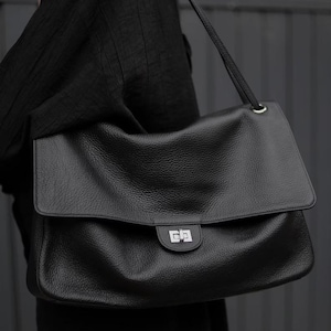 Könnte beinhalten: Eine große schwarze Lederschultertasche mit genarbter Textur, einer breiten Umschlagklappe und einem silberfarbenen rechteckigen Drehverschluss. Die Tasche verfügt über einen dünnen Schulterriemen mit silberfarbenen Beschlägen und das Wort Large.