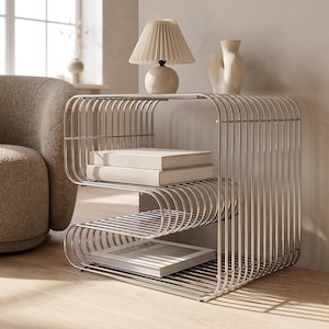 Pode incluir: Uma mesa lateral de metal prateado construída a partir de uma série de hastes curvas paralelas que formam um perfil em S. A mesa possui dois níveis de armazenamento abertos, com dois livros empilhados na prateleira superior e um na inferior.