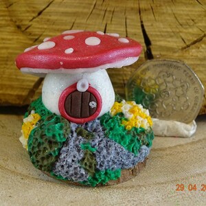 May include: A miniature fairy house made of red and white clay, shaped like a mushroom with a brown door and white glitter accents. The house is surrounded by green and yellow foliage.