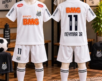 Könnte beinhalten: Ein weißes Fußballtrikot-Set bestehend aus Trikot und Shorts mit schwarzen und orangen Akzenten. Das Trikot zeigt den Text BMG, NETSHOES und CEARA sowie das Santos FC-Wappen. Auf der Rückseite stehen die Nummer 11 und der Name NEYMAR JR in Schwarz.