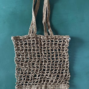May include: A tan-colored tote bag crafted with a crochet net pattern. The bag features a rectangular body with an open-weave design and two matching tan crochet handles attached to the top edge for carrying. The construction is uniform throughout.