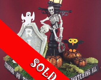 May include: A Halloween-themed centerpiece featuring a skeleton sitting on a black street lamp post with a ghost and a tombstone. The base of the centerpiece is a black tray with the words "Standing on the Watching All" written on it. The tray is decorated with moss, pumpkins, and an owl.