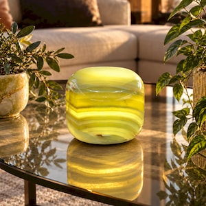 May include: A smooth, rounded, translucent stone object with horizontal bands of light yellow and lime green. The polished surface reflects light, showcasing natural mineral layers and a soft, glowing appearance that highlights the organic texture.