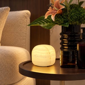 May include: A rounded, cream-colored stone lamp emits a soft, warm glow. It sits on a dark circular table next to a dark brown glass jar, a dark glass pump bottle, and a vase containing green foliage and peach-colored lily flowers.