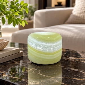 May include: A cylindrical stone object with a smooth, polished surface featuring horizontal bands of translucent light green and white. The stone has a natural, layered appearance with varying shades of pale lime and cream, reflecting light.