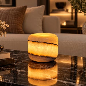 May include: A cylindrical stone lamp with a warm, glowing amber light. The stone features horizontal bands of dark brown and black mineral veins, creating a natural, translucent texture that illuminates the surface beneath it.
