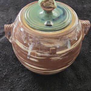 May include: A round ceramic lidded jar with two side handles, featuring a brown and cream swirled glaze with white drip accents. The lid is topped with a rough, light green stone-like finial and has a smooth sage green finish.