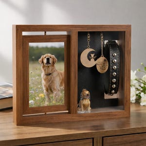 Può includere: Una cornice commemorativa in legno con un portafoto girevole a sinistra contenente la foto di un golden retriever. Il lato destro presenta uno sfondo nero con due ciondoli metallici appesi, una piccola statuetta di cane e un collare nero.