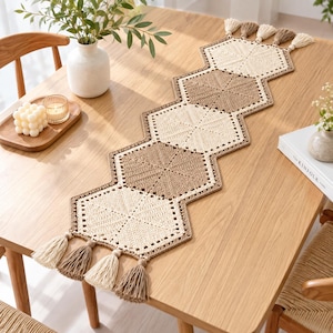 May include: A long, narrow table runner made of crocheted hexagons in alternating cream and tan colours. The runner features a geometric pattern and is finished with decorative tassels in matching cream and tan yarn at both ends.