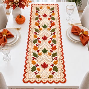 Puede incluir: Un camino de mesa rectangular de ganchillo con un patrón de celosía color crema, decorado con hojas de otoño bordadas en tonos rojo, naranja y verde, acentuado con pequeñas bayas rojas y un borde festoneado de color naranja.