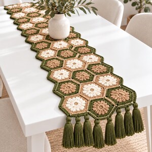 Puede incluir: Un camino de mesa largo hecho de motivos hexagonales de ganchillo en tonos verde oliva, beige y crema. Cada hexágono presenta un patrón floral central. El camino de mesa está rematado con una fila de borlas largas de hilo verde oliva.