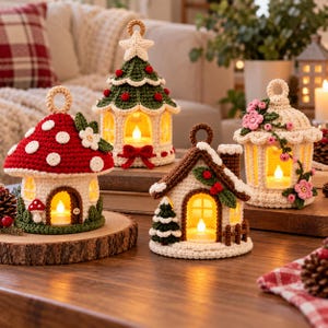 May include: Four crocheted house-shaped lanterns with internal warm LED lights. One is a red mushroom house, one a green Christmas tree, one a snowy cottage, and one a cream house with pink flowers. Each has a top loop for hanging.