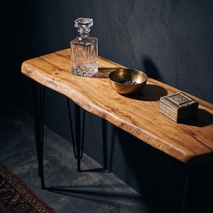 May include: A live edge wooden console table with black metal hairpin legs. On the surface sits a clear glass decanter with a square stopper, a small hammered brass bowl containing dark coffee beans, and a square wooden box with intricate carvings.