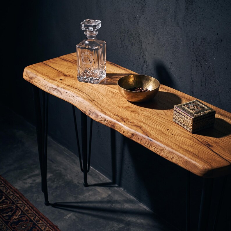 May include: A live edge wooden console table with black metal hairpin legs. On the surface sits a clear glass decanter with a square stopper, a small hammered brass bowl containing dark coffee beans, and a square wooden box with intricate carvings.