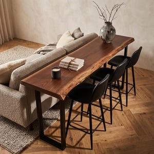 May include: A long, narrow console table with a natural live-edge dark wood top and black metal U-shaped legs. Three black bar stools with curved seats are tucked underneath. A stack of three books and a black candle sit on the table surface.