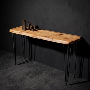May include: A console table featuring a natural live-edge light brown wood slab top with visible grain and knots. The table is supported by four black metal hairpin legs. Three small dark brown geometric decorative objects sit on the surface.