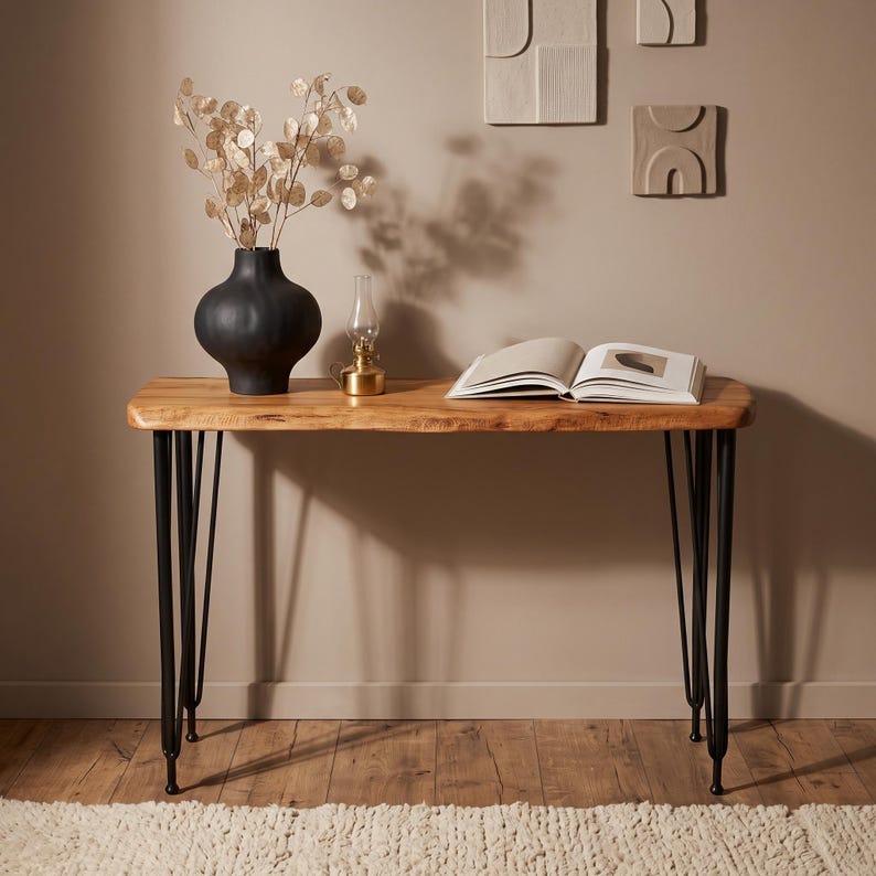 May include: A console table with a natural live-edge wood top and four slender black metal hairpin legs. On the surface sits a matte black rounded vase containing dried silver dollar eucalyptus, a small brass oil lamp, and an open book.