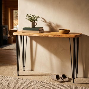May include: A console table with a natural live-edge wood top and four black metal hairpin legs. On the table sits a small white ceramic mug containing a green plant, two stacked rectangular books, and a single round wooden bowl.