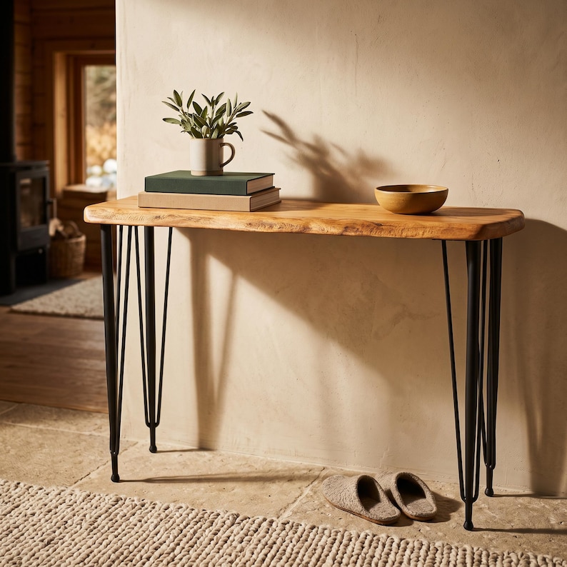 May include: A console table with a natural live-edge wood top and four black metal hairpin legs. On the table sits a small white ceramic mug containing a green plant, two stacked rectangular books, and a single round wooden bowl.