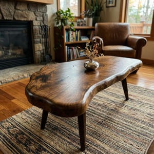 Peut inclure: Une table basse à bord naturel, fabriquée en bois brun foncé avec une finition lisse et polie. La table présente une forme organique irrégulière et repose sur quatre pieds en bois foncé effilés. Un petit vase argenté est posé dessus.