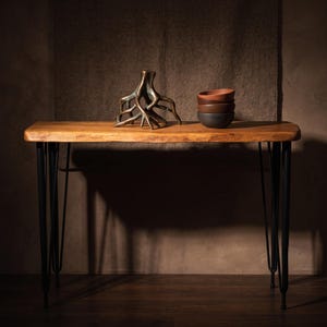 May include: A rectangular wooden console table with a natural live edge top and four black metal hairpin legs. On the table sits a bronze-colored root-shaped sculpture and a stack of three small ceramic bowls in shades of terracotta and dark gray.