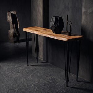 May include: A console table featuring a natural live-edge light brown wooden top supported by four thin black metal hairpin legs. A textured black geometric vase and a tall, slender black metal abstract sculpture sit on the wooden surface.