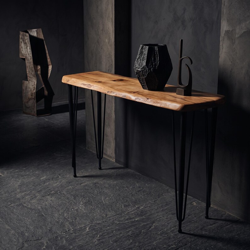May include: A console table featuring a natural live-edge light brown wooden top supported by four thin black metal hairpin legs. A textured black geometric vase and a tall, slender black metal abstract sculpture sit on the wooden surface.