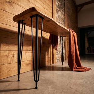 May include: A console table featuring a natural live-edge wooden top with a smooth finish and four black metal hairpin legs. A burnt orange throw blanket with fringed edges is draped over the right side of the wooden surface.