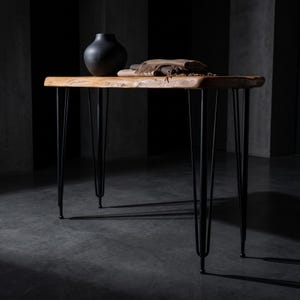 May include: A rectangular wooden table with a natural live edge top and four slender black metal hairpin legs. A matte black rounded vase and a folded brown textile are placed on the surface of the table, which stands on a dark floor.