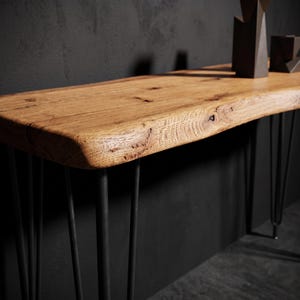May include: A console table featuring a natural live edge wood slab top with visible grain and knots, supported by thin black metal hairpin legs. The surface displays a warm brown tone, contrasting with the dark, slender, angled metal supports.