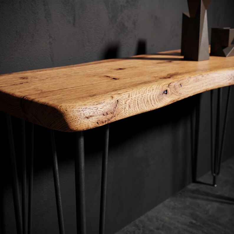 May include: A console table featuring a natural live edge wood slab top with visible grain and knots, supported by thin black metal hairpin legs. The surface displays a warm brown tone, contrasting with the dark, slender, angled metal supports.