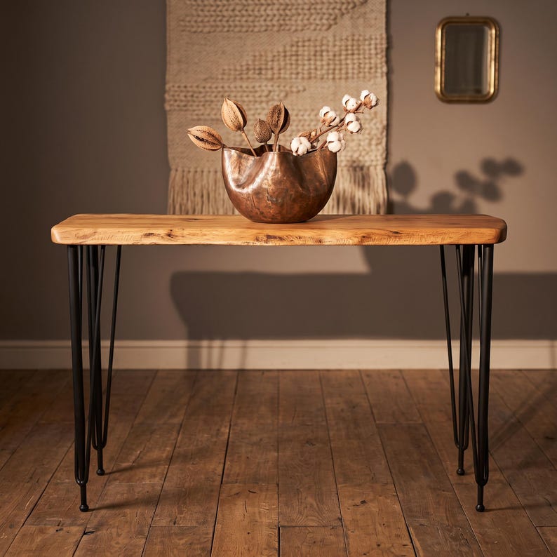 May include: A rectangular wooden console table with a natural live edge top and four black metal hairpin legs. A large, wavy-rimmed copper bowl sits on the table, filled with dried seed pods and white cotton stems, creating a decorative arrangement.