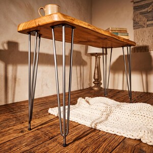 May include: A rectangular wooden desk featuring a natural live edge finish on the tabletop and four sets of slender, silver-toned metal hairpin legs. The desk surface displays a warm, light brown wood grain pattern with visible knots and texture.