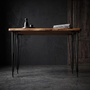 May include: A rectangular wooden table with a natural live edge top and four black metal hairpin legs. On the surface sits a small black bottle, a black cup, and a stack of two books with the visible text Architectural Diary and Luxury Spaces.