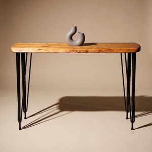 May include: A rectangular console table with a natural light brown wood grain top and four black metal hairpin legs. A small, smooth, matte gray ceramic vase with a curved, abstract shape sits centered on the surface of the wooden table.