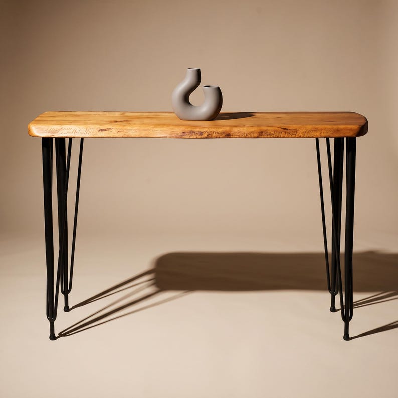 May include: A rectangular console table with a natural light brown wood grain top and four black metal hairpin legs. A small, smooth, matte gray ceramic vase with a curved, abstract shape sits centered on the surface of the wooden table.