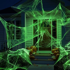 May include: A house exterior decorated with bright green artificial spider webs draped over the front porch, bushes, and steps. Several orange pumpkins of varying sizes are arranged on the steps and near the entrance of the home.