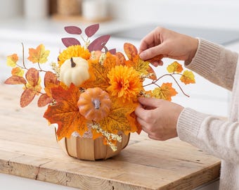 May include: A decorative arrangement featuring artificial pumpkins, orange dahlia flowers, and autumn-coloured maple leaves in a small wooden basket. The display includes white and orange pumpkins nestled among foliage with yellow, red, and brown tones.