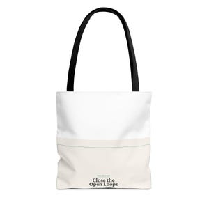 May include: A white and cream-colored tote bag with black shoulder straps. The bag features a horizontal line across the middle and the text "Close the Open Loops" printed in black at the bottom center, with smaller text above it.