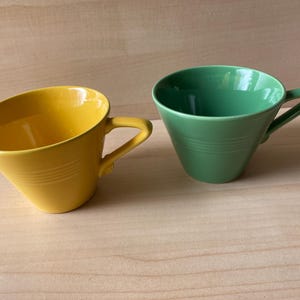 May include: Two ceramic teacups, one yellow and one green, sit side-by-side. Each cup has a handle and a slanted design with horizontal grooves. The cups are on a light brown wooden surface.