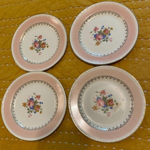 May include: Four white ceramic plates with a pink border and gold trim. Each plate features a floral bouquet in the center with pink, blue, and yellow flowers. The plates are arranged on a yellow surface.