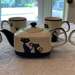 May include: A cream-colored tea set with black cat and paw print designs. The set includes a teapot with a lid, and four mugs. The teapot and two mugs feature a silhouette of two cats. The other two mugs have paw prints.