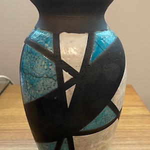 May include: A decorative ceramic vase with a black rim and a geometric design. The vase features a textured surface with sections of white, teal, and black. The vase is approximately 10 inches tall.