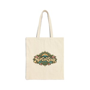 Peut inclure: Un sac fourre-tout en toile couleur crème arborant un motif central avec le texte NorCal Soak. L'illustration comprend un sommet de montagne, de l'eau bleue et des éléments floraux disposés en un motif décoratif symétrique de type mandala.